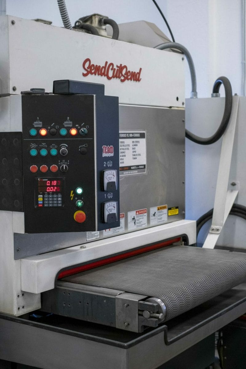 Image shows a wet deburring process machine with the logo "SendCutSend" in red above the conveyor belt.
