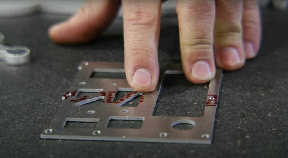 Screenshot from the video showing Jake pointing to red manufacturing markings left on a sheet metal part prior to deburring.