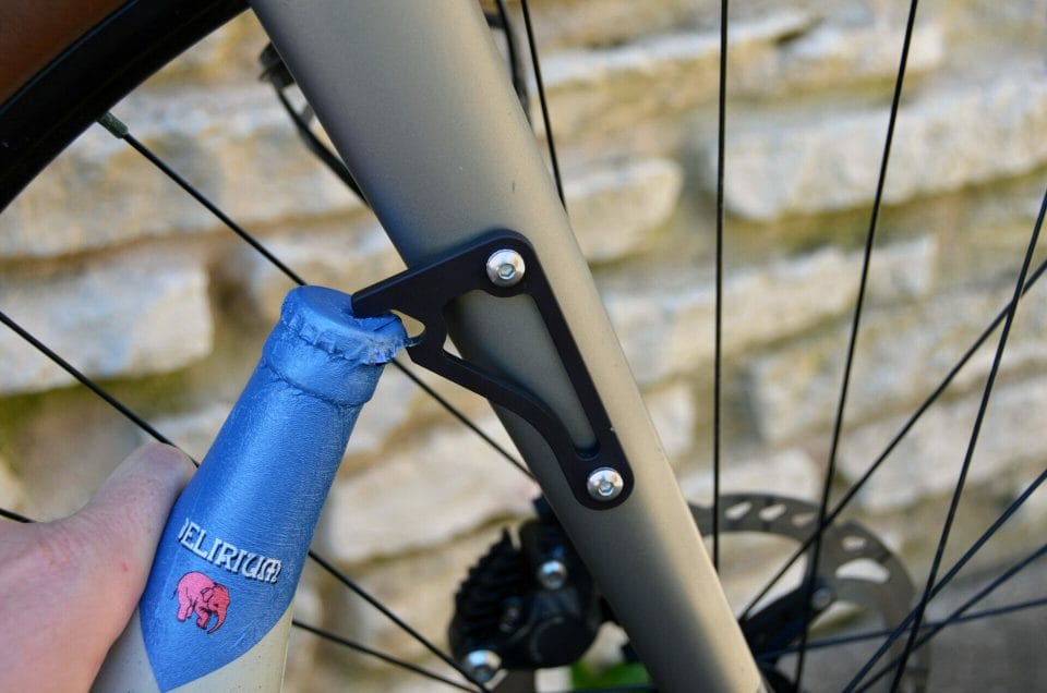 A black anodized aluminum bottle lever from Foothill Products mounted on a bike, with someone opening a blue topped Delirium beer.