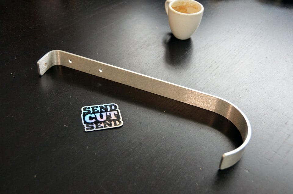 A coining bent aluminum mounting hook resting on a black table next to a SendCutSend holographic sticker and an empty espresso cup.