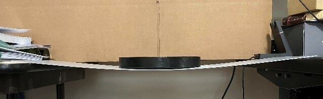 A black weight on the flat sheet metal part, causing it to bend in the center.